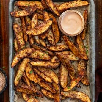 Sheet pan with Crispy Potato Wedges and dipping sauce on it on a wooden table from overhead.