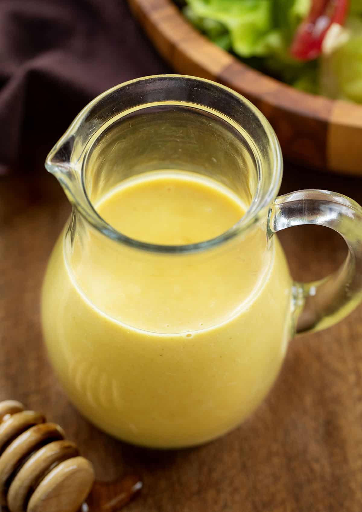 Small pitcher of Honey Mustard Dressing on a wooden table with honey dipper and fresh salad.