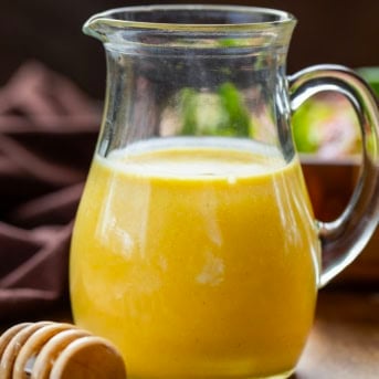 Small pitcher of Honey Mustard Dressing on a wooden table with honey dipper and fresh salad.