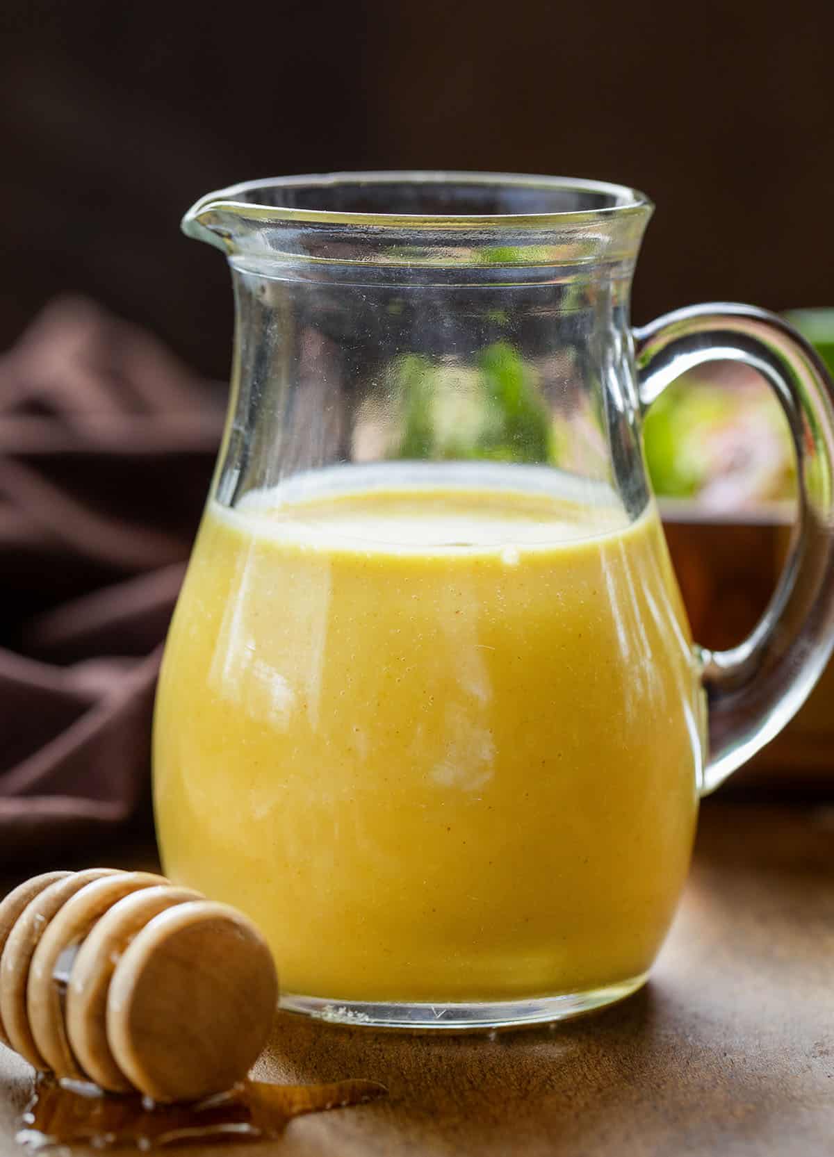 Small pitcher of Honey Mustard Dressing on a wooden table with honey dipper and fresh salad.