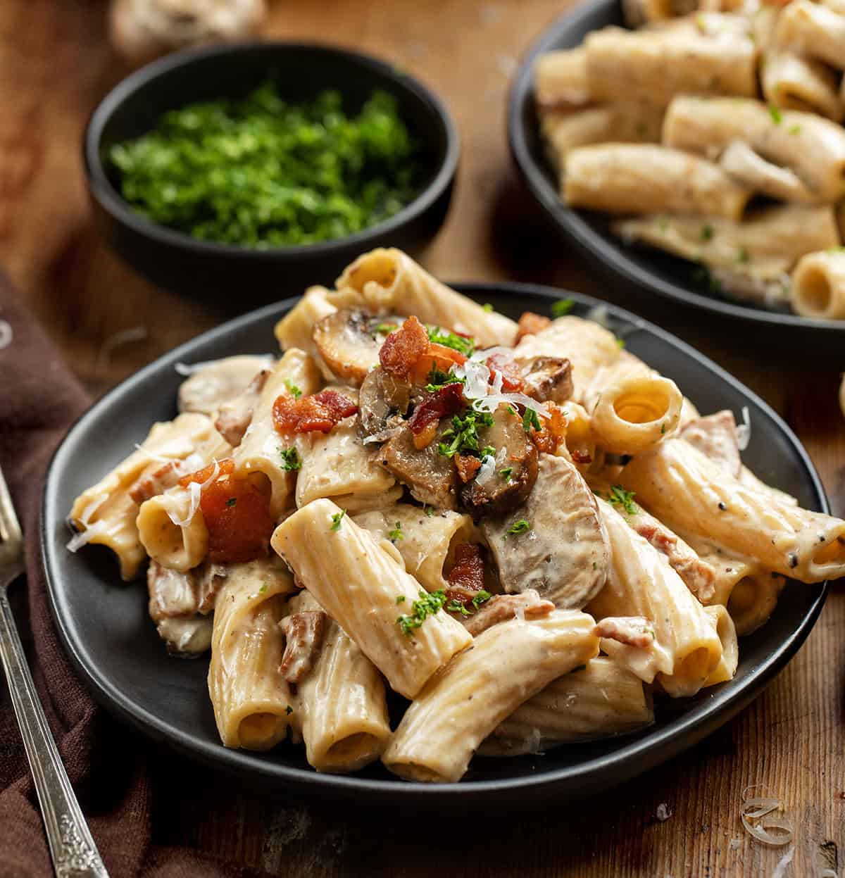 This mushroom and bacon rigatoni is a rich, creamy pasta made with crispy bacon, sautéed mushrooms, and a silky Parmesan cream sauce. It’s hearty, comforting, and full of deep savory flavor, with a touch of lemon to keep it balanced. Black plates of Mushroom and Bacon Rigatoni on a wooden table close up.