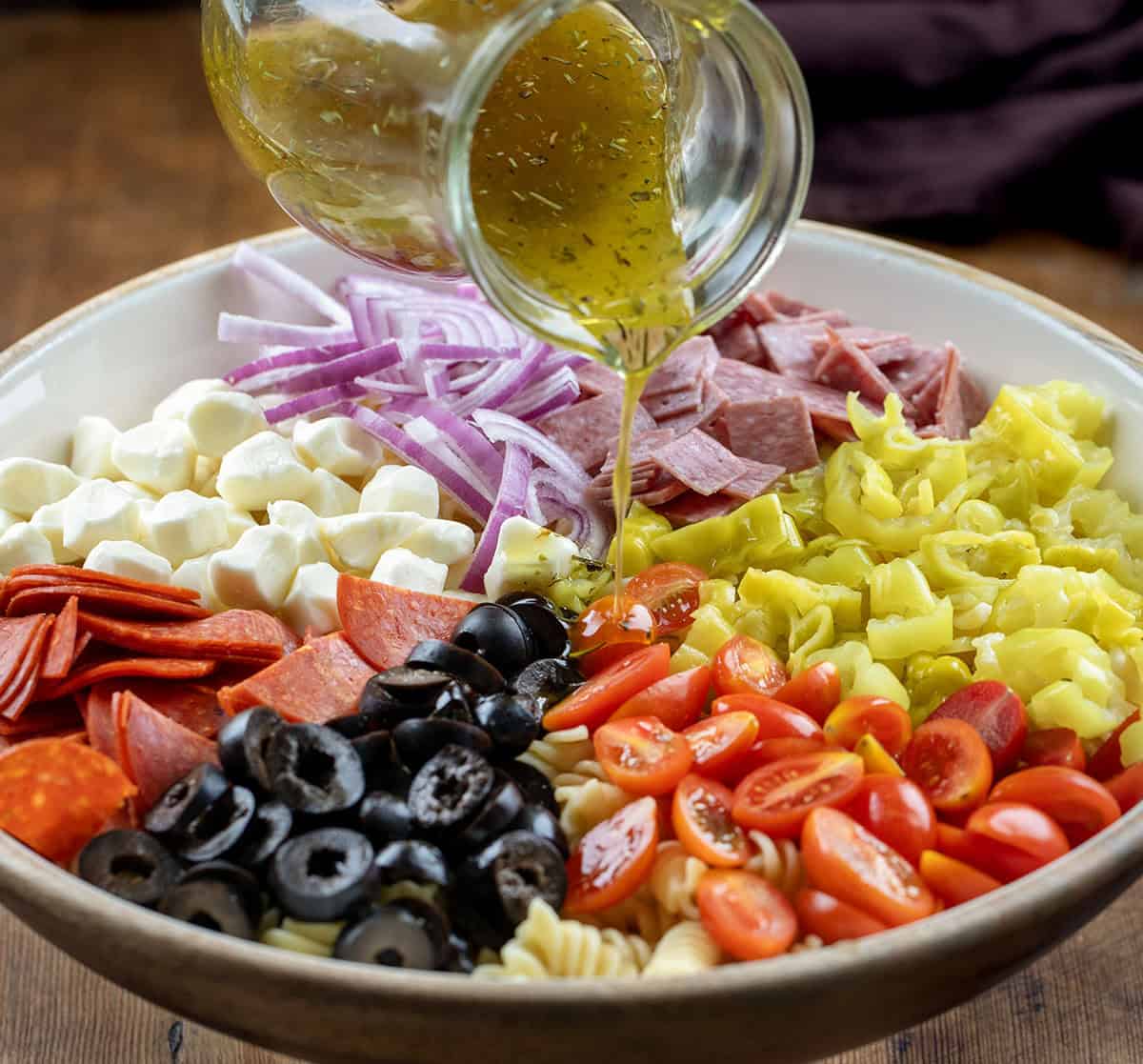 This Pepperoncini Pasta Salad is bold, tangy, and packed with all the good stuff. Tender rotini is tossed with mozzarella, pepperoni, salami, fresh veggies, and plenty of pepperoncini for that zesty bite in every forkful! Pouring dressing over Pepperoncini Pasta Salad ingredients.