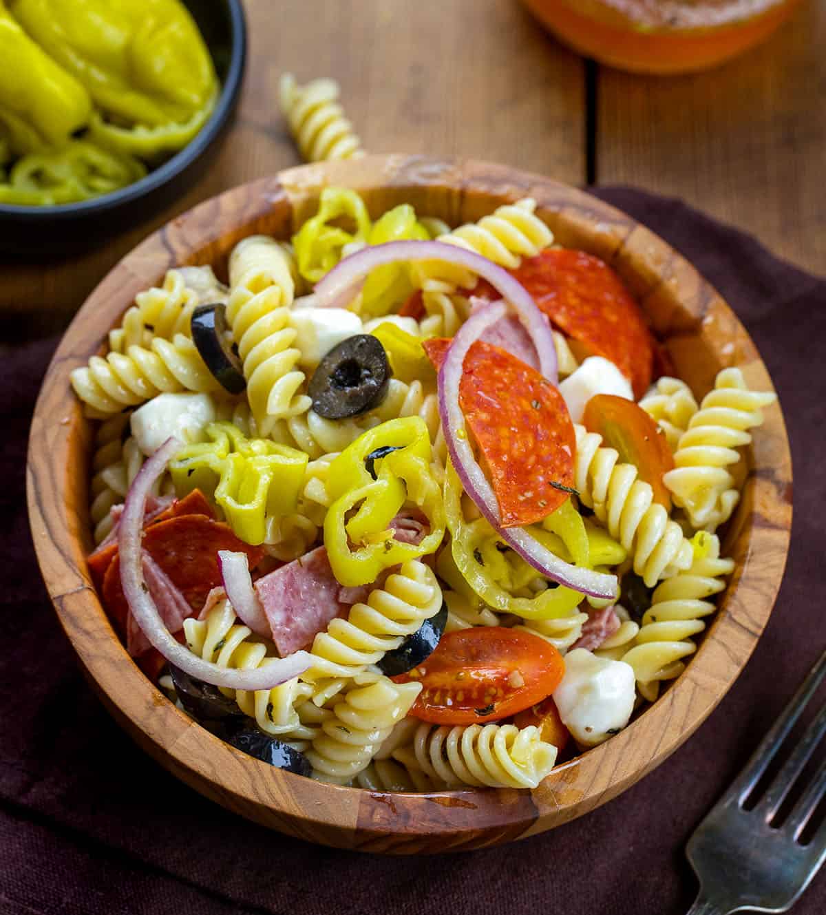 This Pepperoncini Pasta Salad is bold, tangy, and packed with all the good stuff. Tender rotini is tossed with mozzarella, pepperoni, salami, fresh veggies, and plenty of pepperoncini for that zesty bite in every forkful! Wooden bowl of Pepperoncini Pasta Salad on a wooden table.