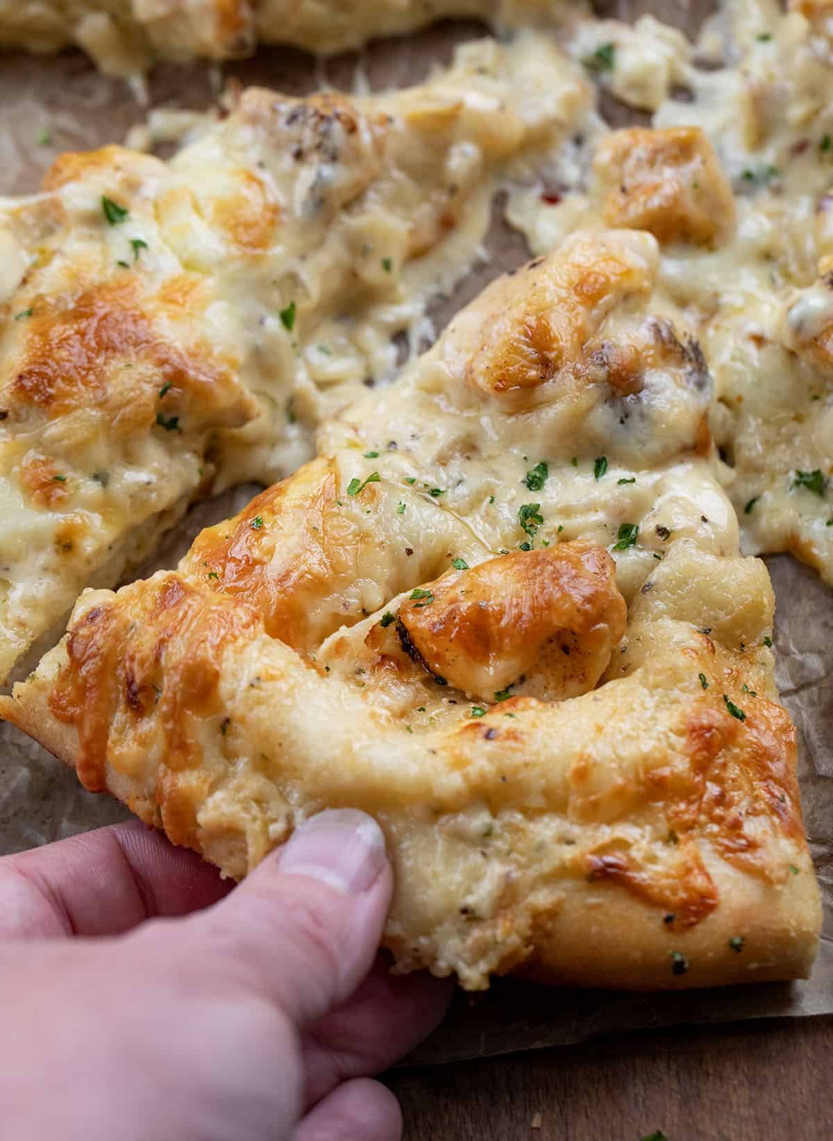 I loved my Chicken Alfredo Garlic Bread so much, I had to make this Chicken Alfredo Pizza, too! It takes everything you love about creamy Alfredo sauce, tender chicken, and melty cheese, and piles it all onto a soft homemade pizza dough that bakes up golden and irresistible! Hand picking up a piece of Chicken Alfredo Pizza showing the crispy crust.