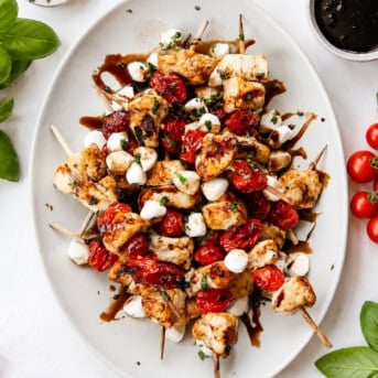 Platter of Chicken Caprese Skewers on a white table with tomatoes and basil.