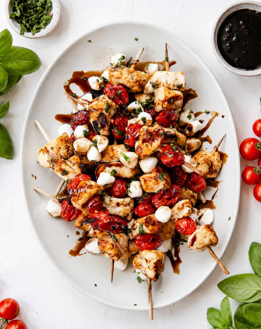 Platter of Chicken Caprese Skewers on a white table with tomatoes and basil Platter of Chicken Caprese Skewers on a white table with tomatoes and basil.