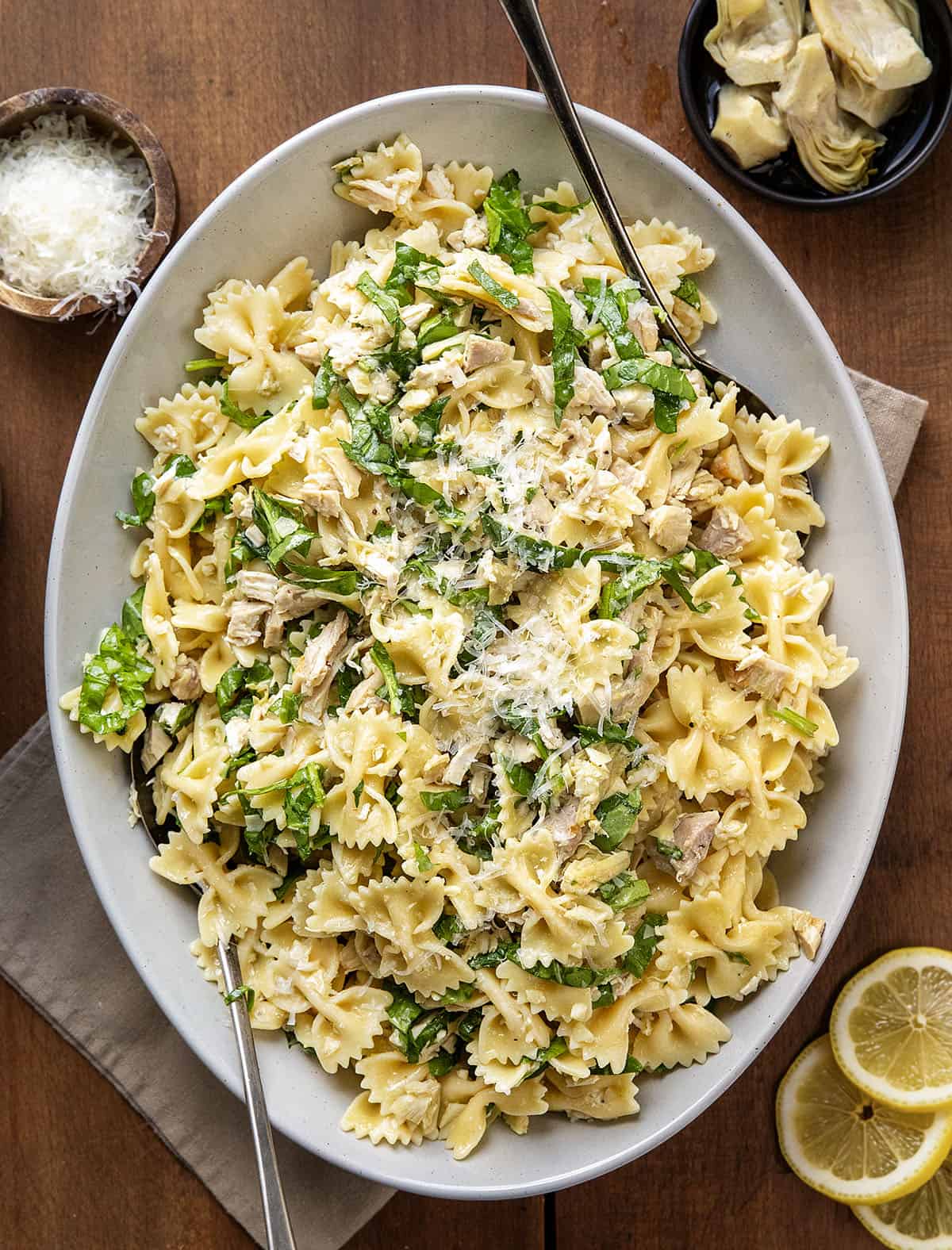 This Lemon Parmesan Pasta Salad with Chicken is a light, fresh pasta salad made with tender chicken, pasta, parmesan, and a bright lemon-garlic vinaigrette! Serving platter of Chicken Lemon Parmesan Pasta Salad with serving spoons on a wooden table from overhead.