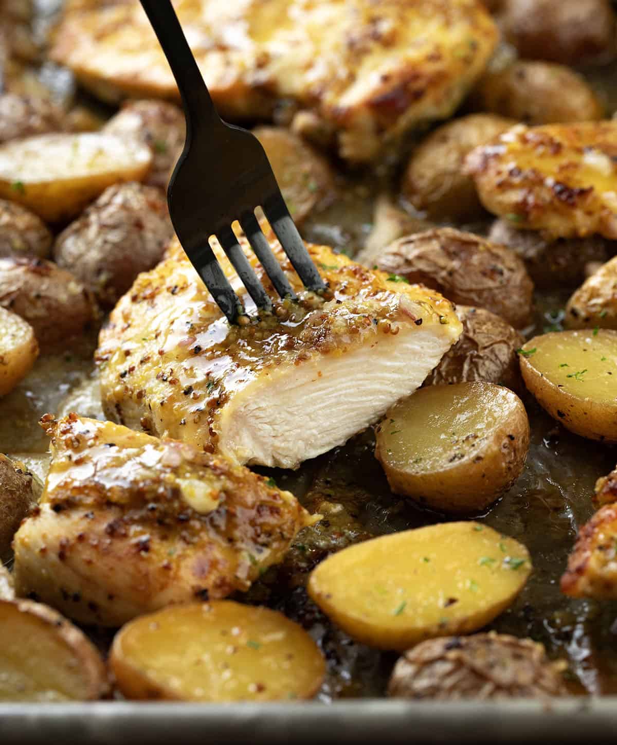 This One Pan Honey Mustard Chicken and Potatoes is an easy sheet pan dinner made with juicy marinated chicken, crispy roasted potatoes, and a sweet-tangy honey mustard glaze! Halved piece of Honey Mustard Chicken and Potatoes on the sheet pan with potatoes.
