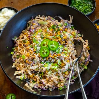 Black bowl filled with Mexican Street Corn Slaw and serving spoons on a wooden table.