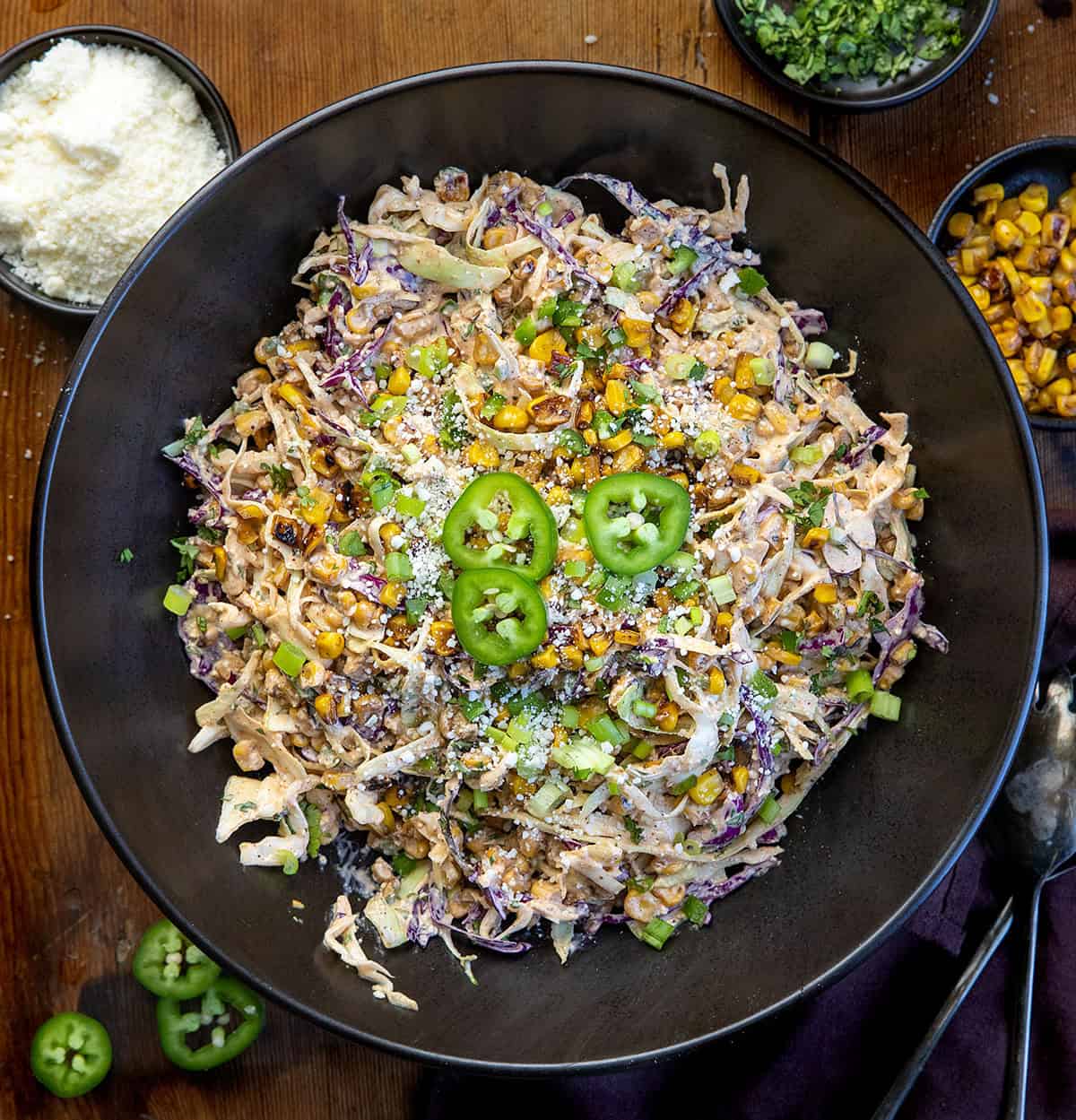 This Mexican Street Corn Slaw is a fresh, crunchy side dish inspired by classic elote. It’s made with shredded cabbage, sweet corn, a creamy lime dressing, and just the right amount of spice! Black bowl filled with Mexican Street Corn Slaw on a wooden table from overhead.