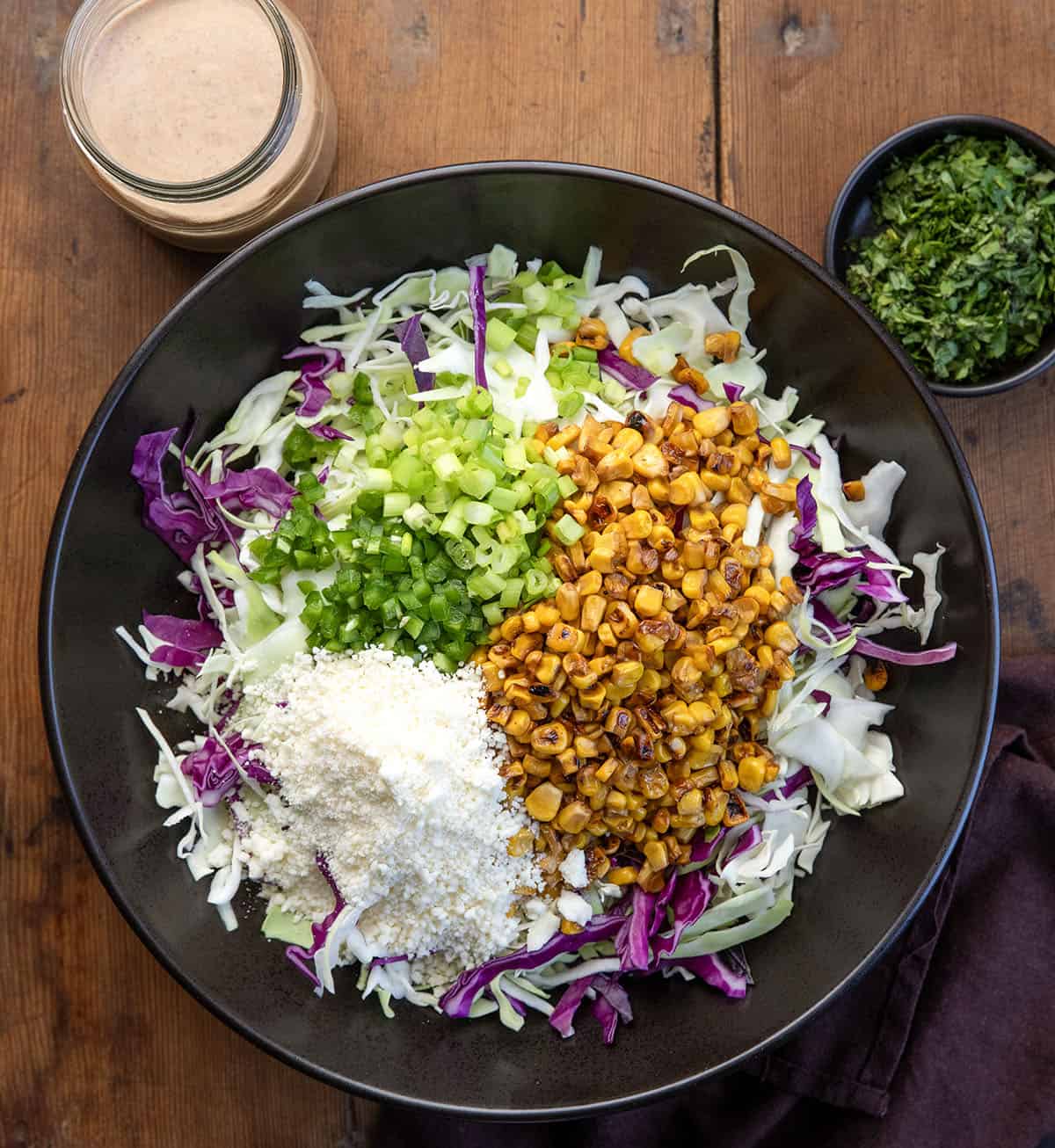 This Mexican Street Corn Slaw is a fresh, crunchy side dish inspired by classic elote. It’s made with shredded cabbage, sweet corn, a creamy lime dressing, and just the right amount of spice! Raw ingredients for Mexican Street Corn Slaw in a black bowl on a wooden table from overhead.