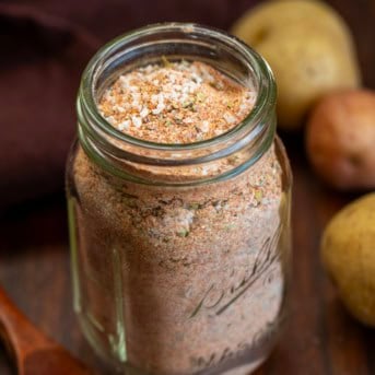 Jar of Potato Seasoning on a wooden table with a small measuring spoon and potatoes.