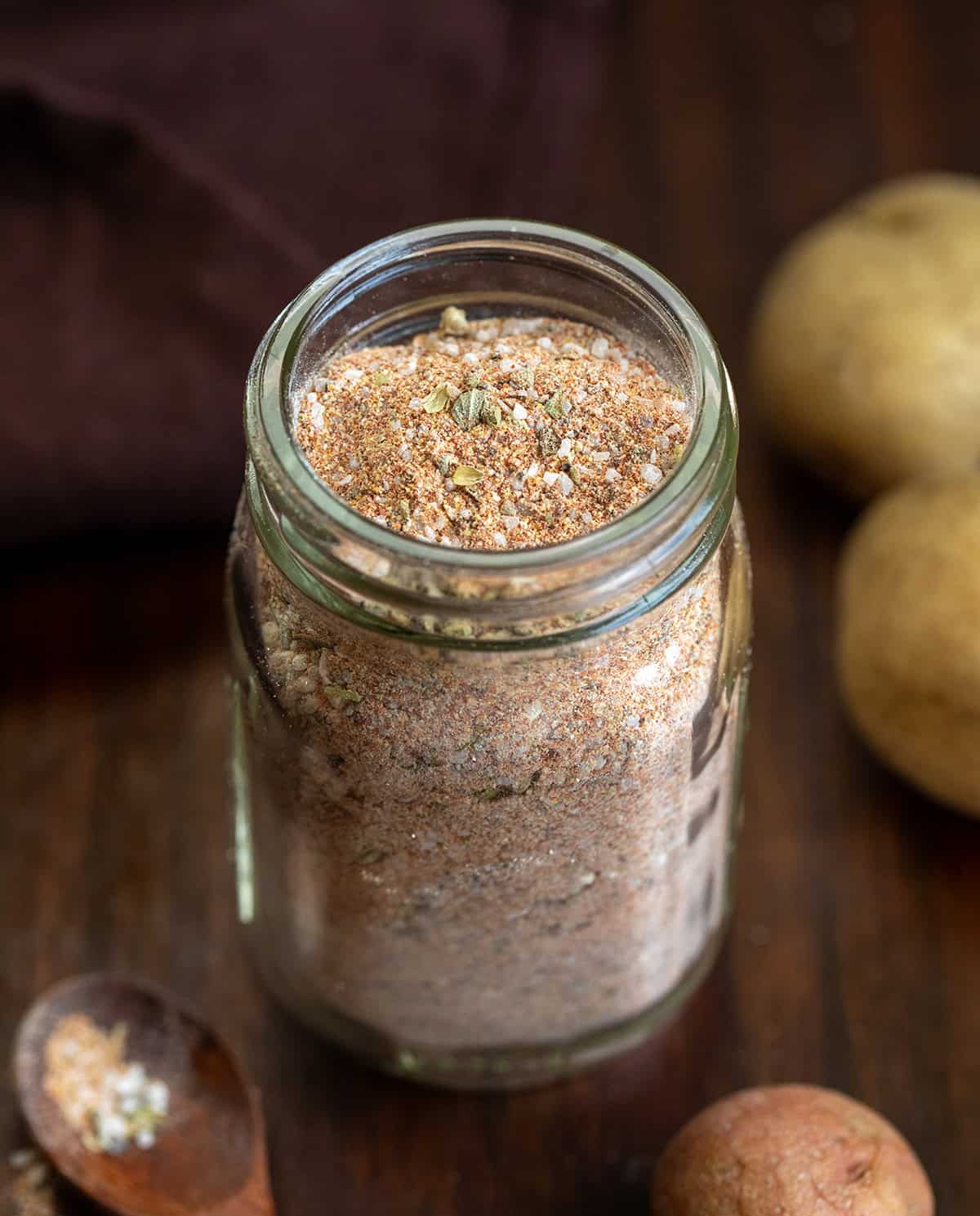 This homemade Potato Seasoning is a simple, savory blend that takes roasted, baked, or air-fried potatoes to the next level! Jar of Potato Seasoning on a wooden table with a small measuring spoon and potatoes.