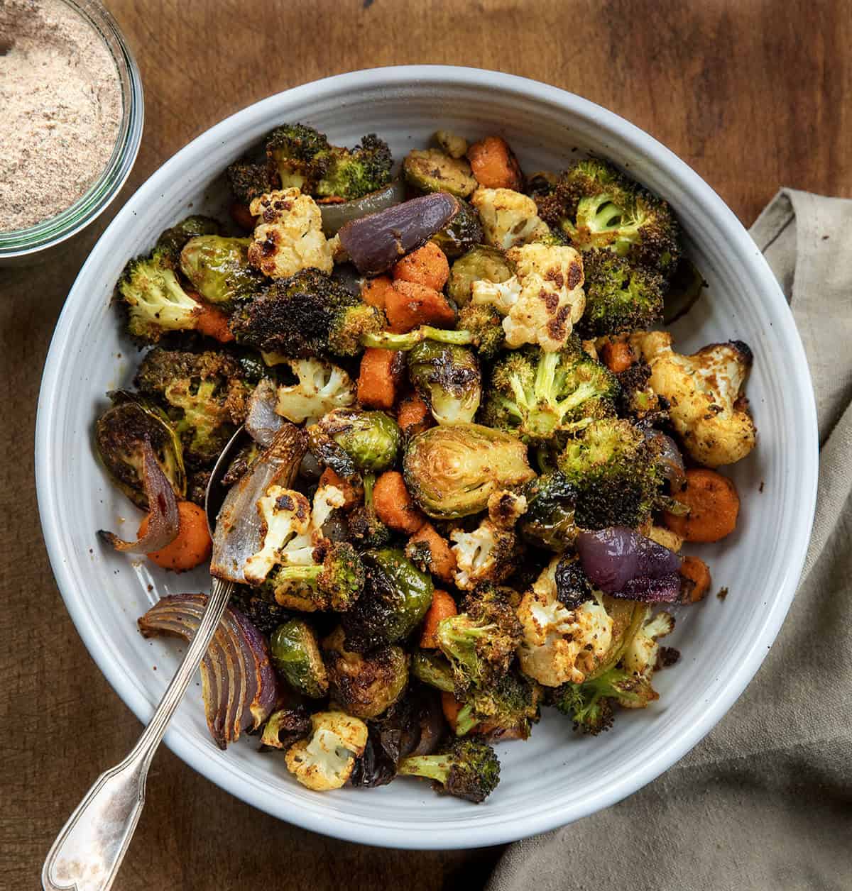 These Sheet Pan Roasted Vegetables are an easy, flavorful side dish made with a colorful mix of broccoli, Brussels sprouts, carrots, cauliflower, and red onion! Bowl of Sheet Pan Roasted Vegetables on a wooden table with a jar of vegetable seasoning.