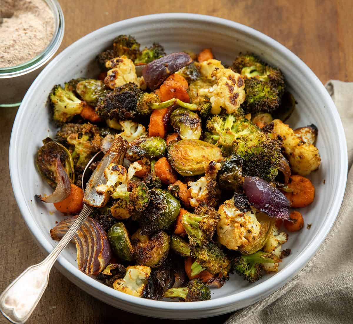 These Sheet Pan Roasted Vegetables are an easy, flavorful side dish made with a colorful mix of broccoli, Brussels sprouts, carrots, cauliflower, and red onion! Bowl of Sheet Pan Roasted Vegetables on a wooden table with a jar of vegetable seasoning.