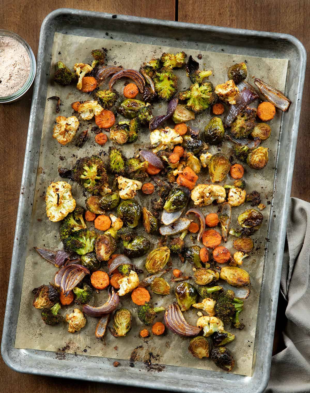 These Sheet Pan Roasted Vegetables are an easy, flavorful side dish made with a colorful mix of broccoli, Brussels sprouts, carrots, cauliflower, and red onion! Sheet Pan Roasted Vegetables on a wooden table with a jar of vegetable seasoning from overhead.