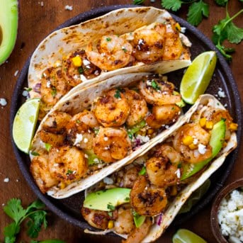 Plate of Shrimp Tacos with Mexican Street Corn Slaw on a wooden table with lime wedges and avocado.