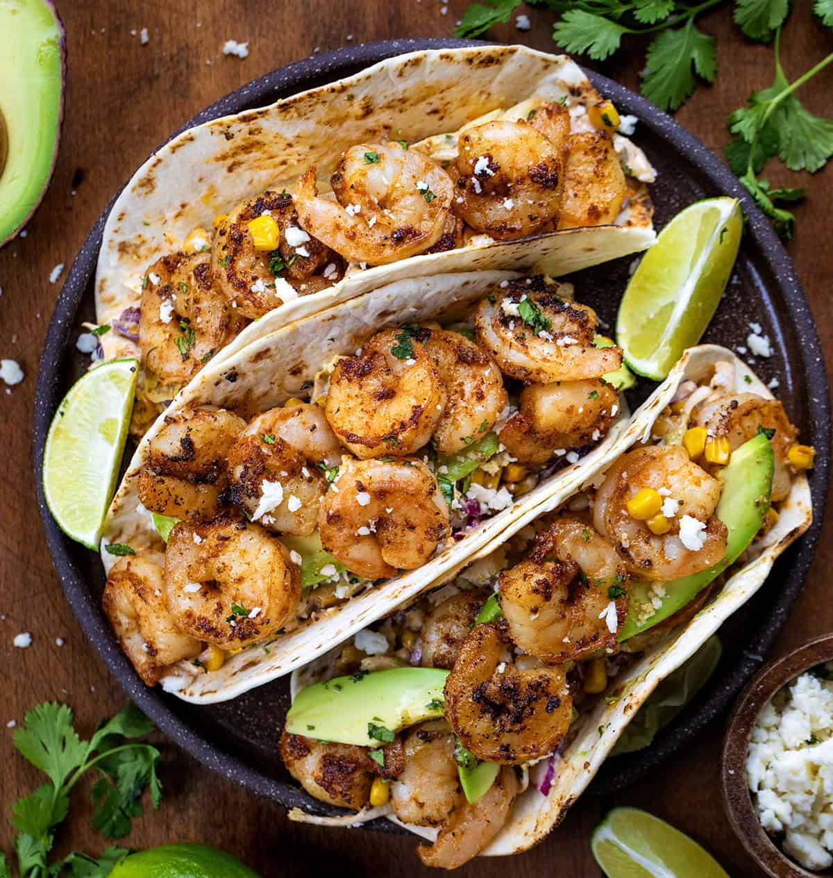 Plate of Shrimp Tacos with Mexican Street Corn Slaw on a wooden table with lime wedges and avocado.