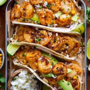 Tray of Shrimp Tacos with Mexican Street Corn Slaw on a wooden table with roasted corn, avocado, and fresh lime.