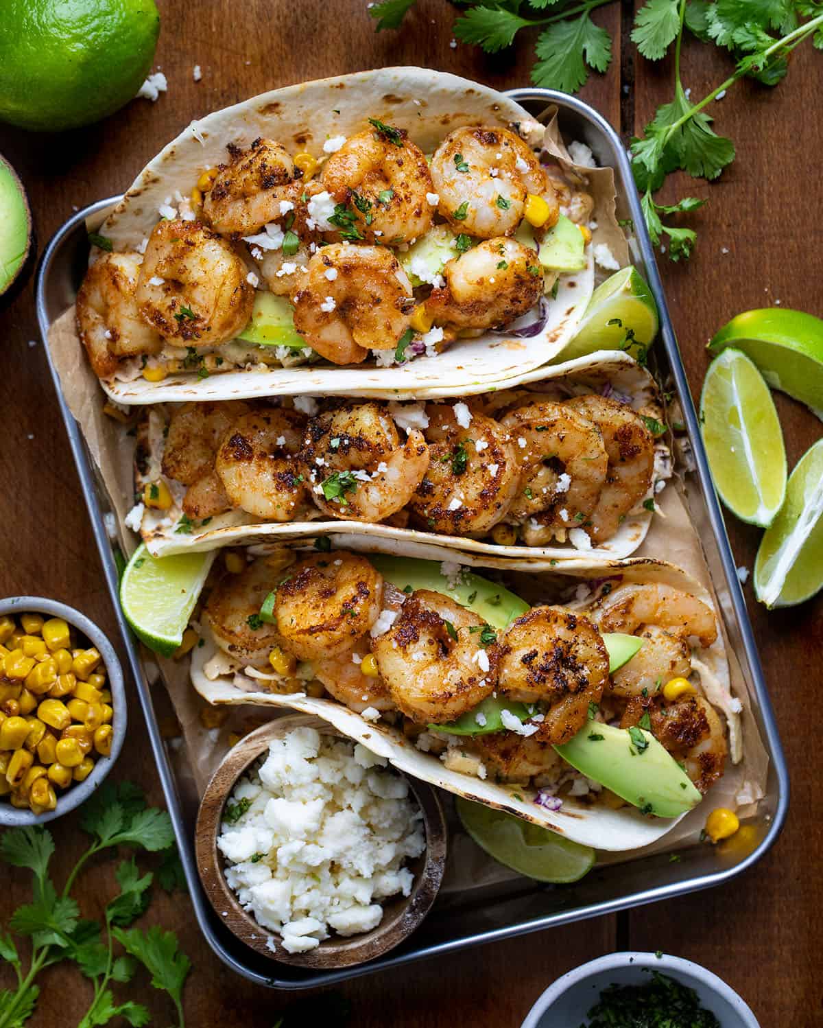 Tray of Shrimp Tacos with Mexican Street Corn Slaw on a wooden table with roasted corn, avocado, and fresh lime.