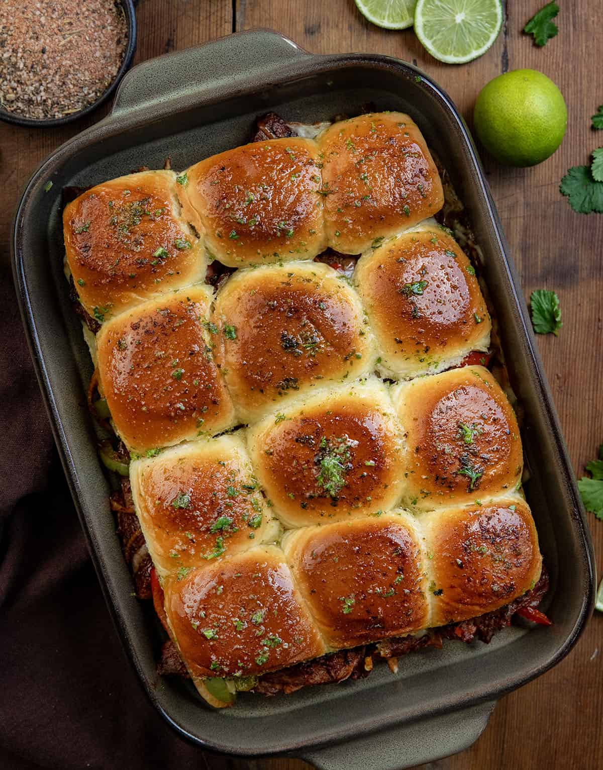 These Steak Fajita Sliders are loaded with tender, seasoned steak, sautéed peppers and onions, and melty cheese all baked inside soft Hawaiian rolls! Pan of Steak Fajita Sliders on a wooden table from overhead with limes and homemade steak seasoning.