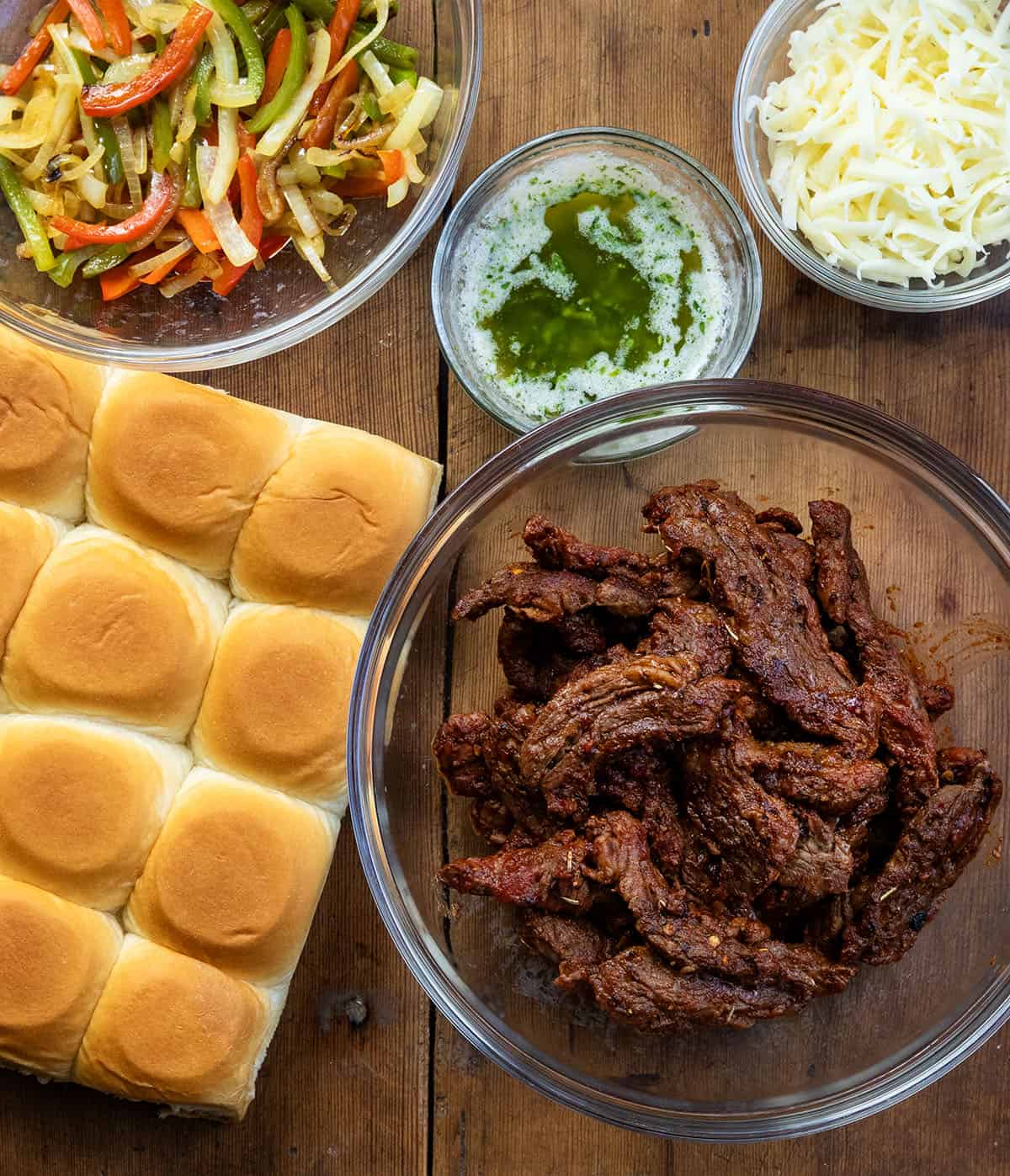 These Steak Fajita Sliders are loaded with tender, seasoned steak, sautéed peppers and onions, and melty cheese all baked inside soft Hawaiian rolls! Ingredients for Steak Fajita Sliders on a wooden table from overhead.