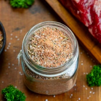 Glass jar of Steak Seasoning on a wooden table next to a steak.