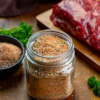 Glass jar of Steak Seasoning on a wooden table next to a steak.