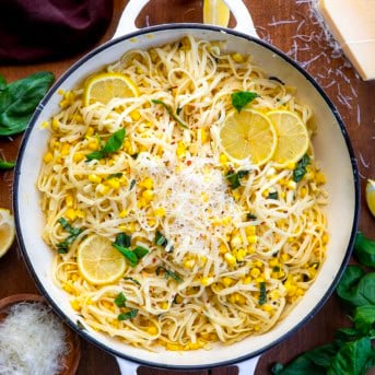 Skillet of Summer Lemon Corn Pasta on a wooden table from overhead surrounded by fresh parmesan, lemon, and basil.