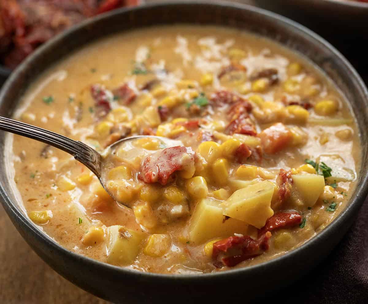This Sun-Dried Tomato Corn Chowder is a rich, creamy soup made with sweet corn, tender vegetables, smoky bacon, and sun-dried tomatoes for extra depth of flavor! Bowl of Sun-Dried Tomato Corn Chowder on a wooden table with a spoon picking up a bite.