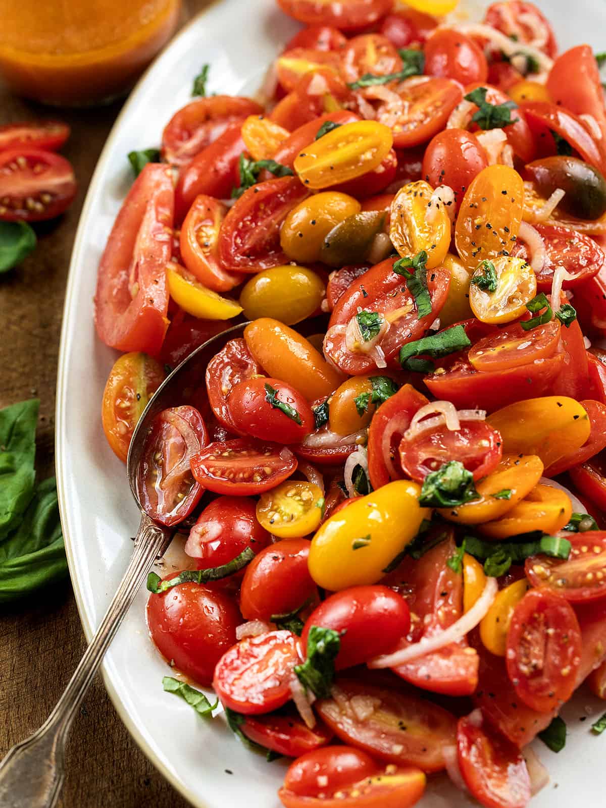 This Simple Tomato Salad is the easiest way to make the most of ripe, in-season tomatoes. Juicy wedges and sweet cherry tomatoes are tossed with shallot, garlic, fresh basil, and a bright vinaigrette made from tomato juices, lemon, and red wine vinegar! Close up of Tomato Salad on a wooden table with a spoon on the platter.