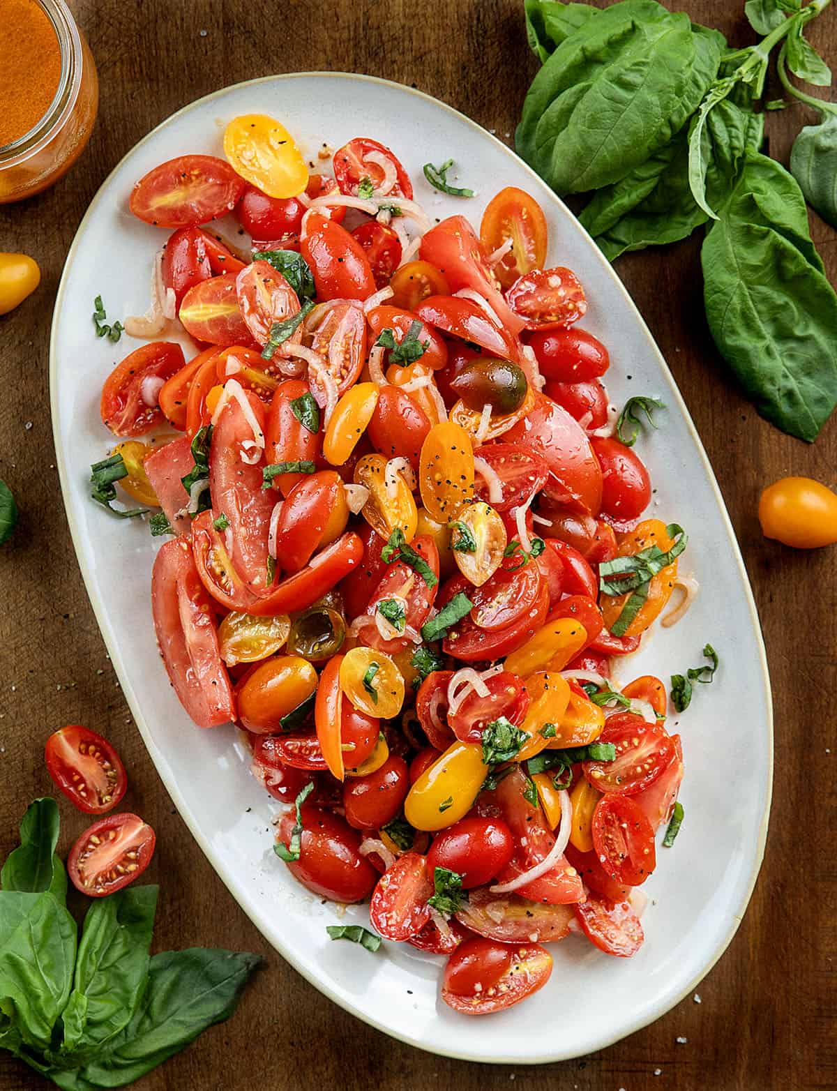 This Simple Tomato Salad is the easiest way to make the most of ripe, in-season tomatoes. Juicy wedges and sweet cherry tomatoes are tossed with shallot, garlic, fresh basil, and a bright vinaigrette made from tomato juices, lemon, and red wine vinegar! Platter of Tomato Salad on a wooden table with fresh tomatoes and basil around the dish.