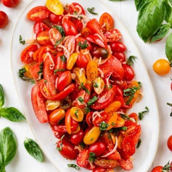 White platter of Tomato Salad on a white table surrounded by fresh tomatoes and basil from overhead.