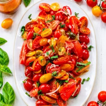 White platter of Tomato Salad on a white table surrounded by fresh tomatoes and basil from overhead.