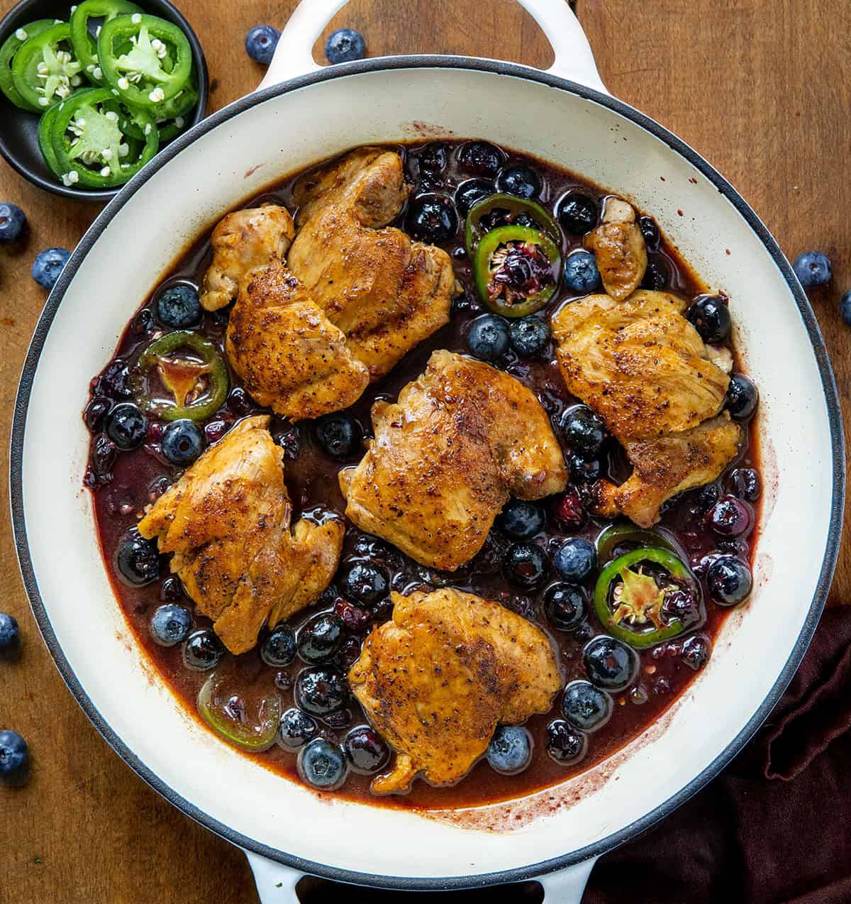 White skillet of Blueberry Jalapeño Chicken on a wooden table with fresh jalapeno and blueberries White skillet of Blueberry Jalapeño Chicken on a wooden table with fresh jalapeno and blueberries.