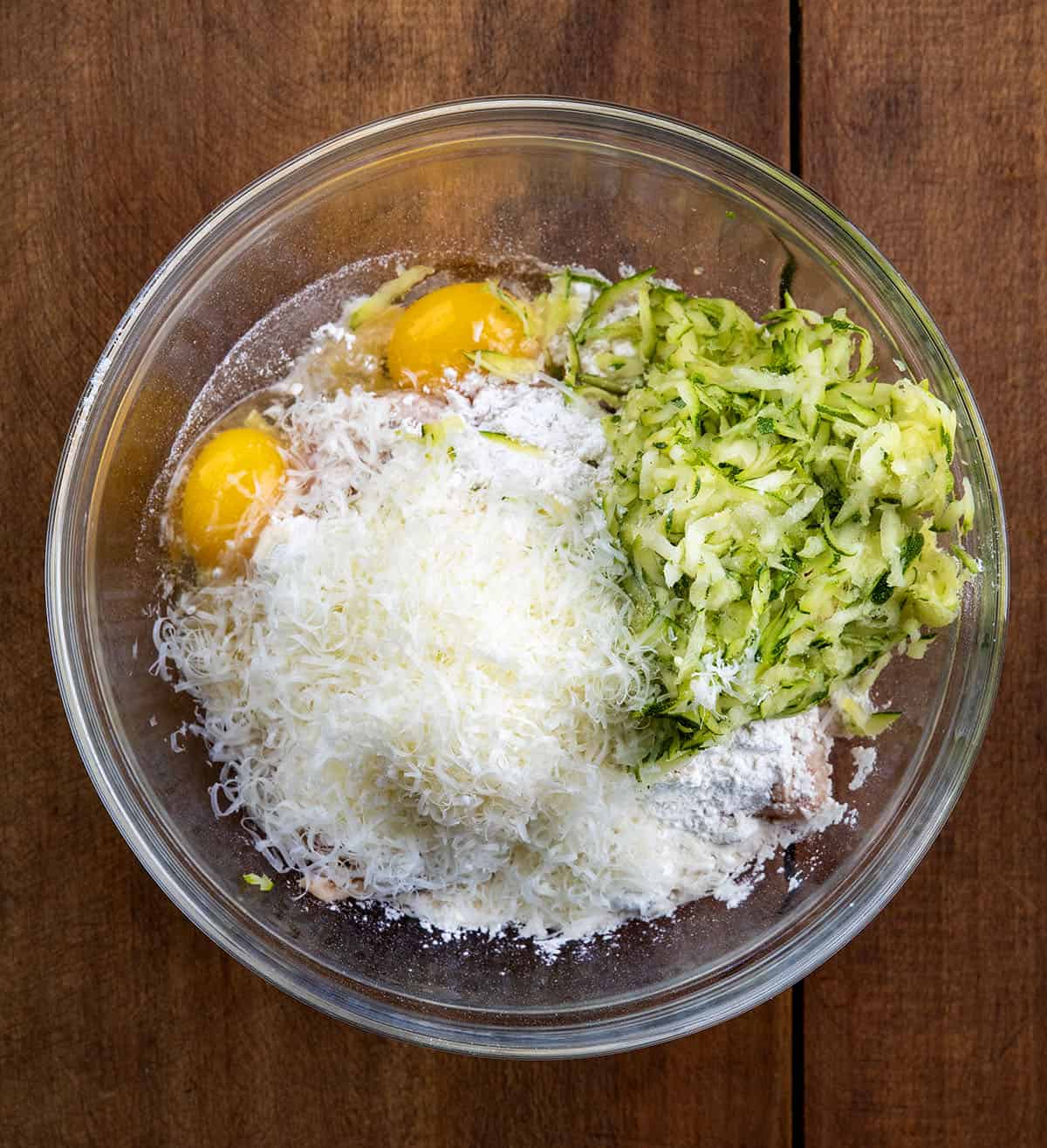 Raw ingredients for Cheesy Zucchini Chicken Fritters in a glass bowl on a wooden table.