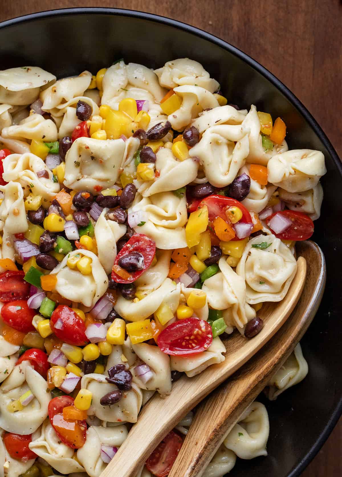 Close up of Cowboy Caviar Tortellini Salad in a black bowl with wooden spoons Close up of Cowboy Caviar Tortellini Salad in a black bowl with wooden spoons.