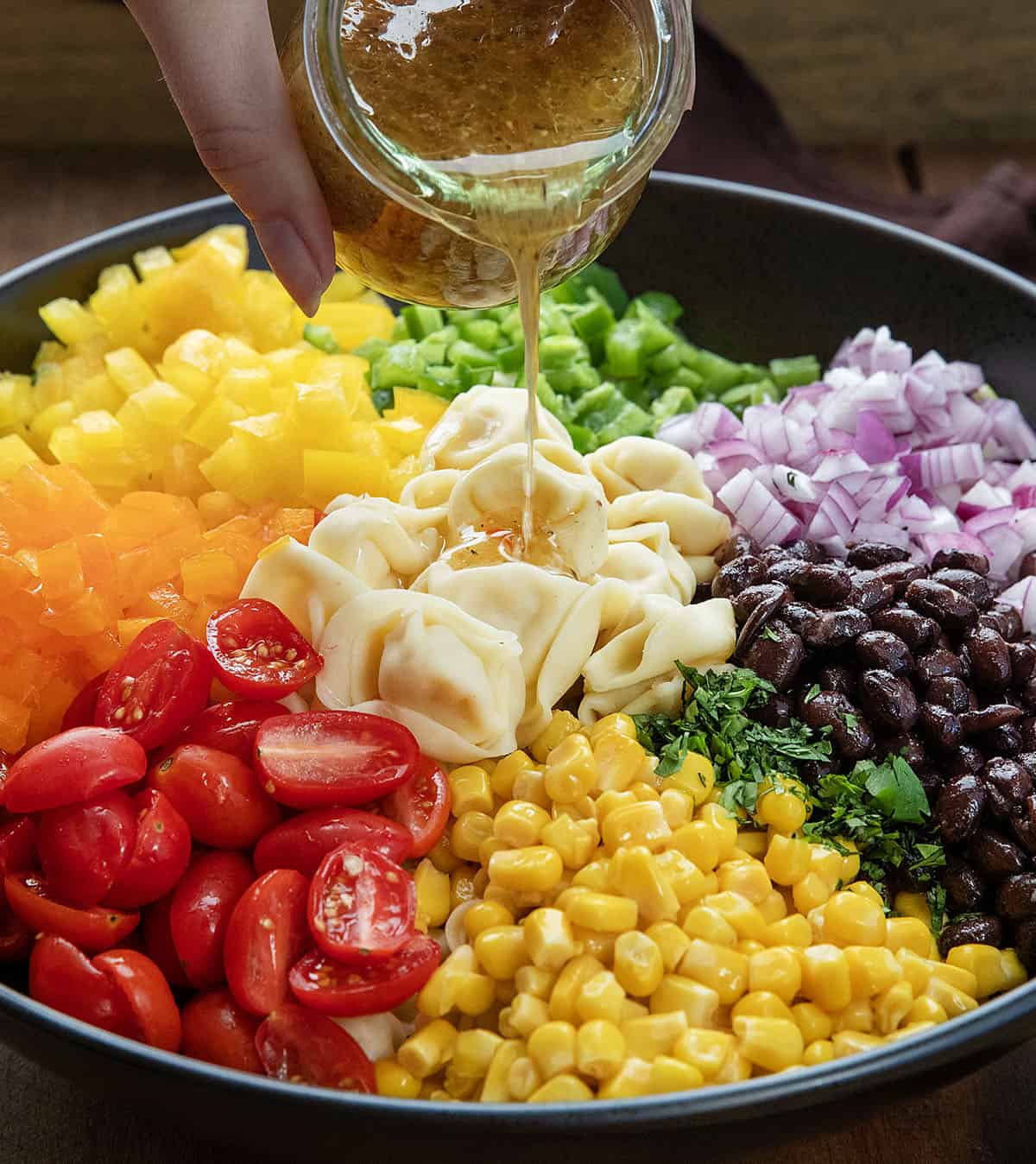 Pouring dressing over the unmixed ingredients for Cowboy Caviar Tortellini Salad in a black bowl Pouring dressing over the unmixed ingredients for Cowboy Caviar Tortellini Salad in a black bowl.