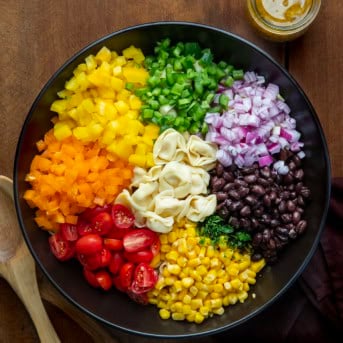 Raw ingredients for Cowboy Caviar Tortellini Salad in a black bowl, showing peppers, jalapeños, corn, beans, tortellini, onion, tomato, and a jar of viniagrette.