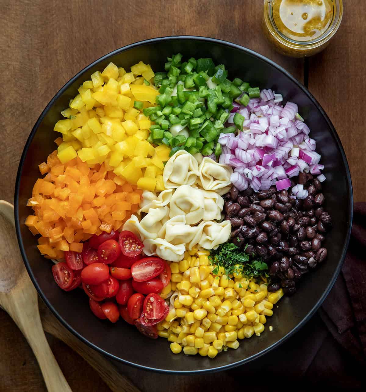 Raw ingredients for Cowboy Caviar Tortellini Salad in a black bowl, showing peppers, jalapeños, corn, beans, tortellini, onion, tomato, and a jar of viniagrette Raw ingredients for Cowboy Caviar Tortellini Salad in a black bowl, showing peppers, jalapeños, corn, beans, tortellini, onion, tomato, and a jar of viniagrette.