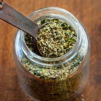 Glass jar of Italian Seasoning with a measuring spoon in the jar on a wooden table.