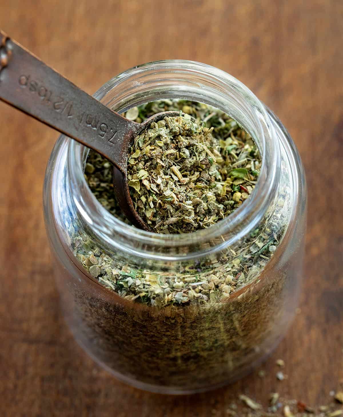 Glass jar of Italian Seasoning with a measuring spoon in the jar on a wooden table Glass jar of Italian Seasoning with a measuring spoon in the jar on a wooden table.