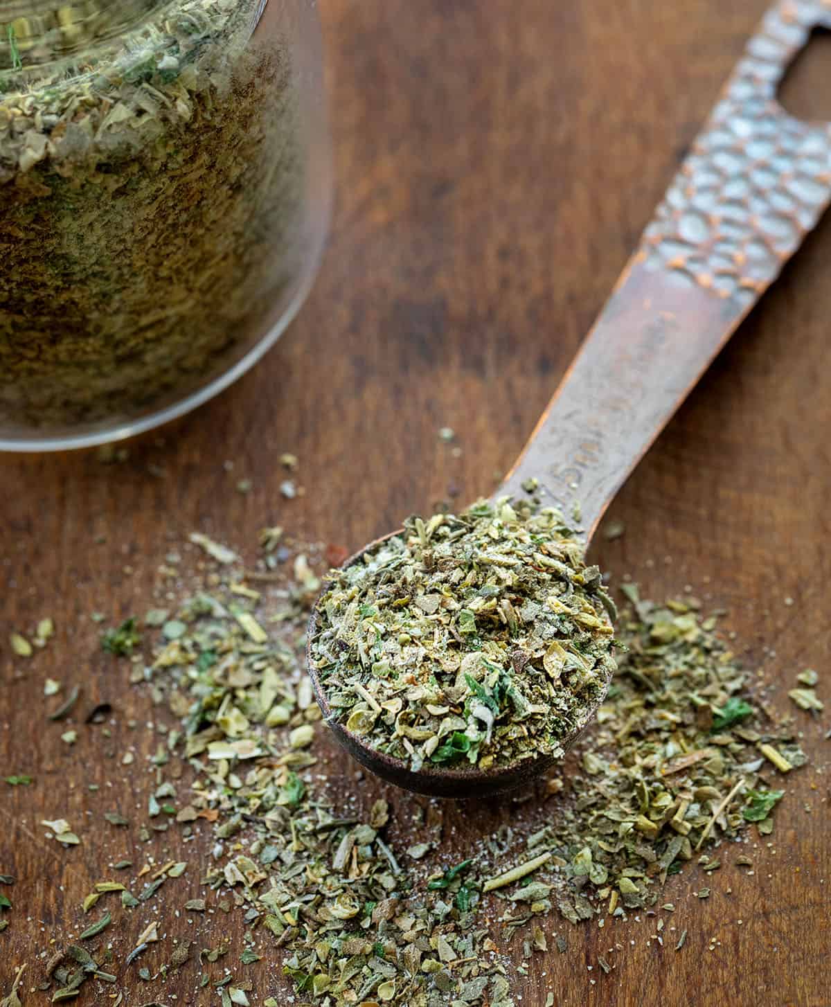 Measuring spoon filled with Italian Seasoning on a wooden table next to the jar Measuring spoon filled with Italian Seasoning on a wooden table next to the jar.