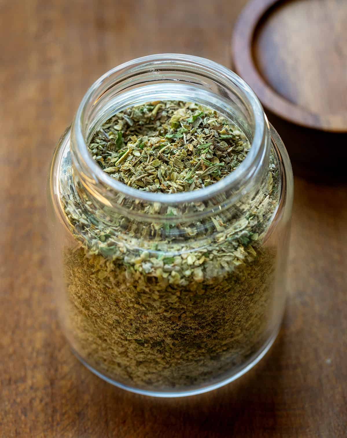 Glass jar of homemade Italian Seasoning with the lid off to the side on a wooden table Glass jar of homemade Italian Seasoning with the lid off to the side on a wooden table.