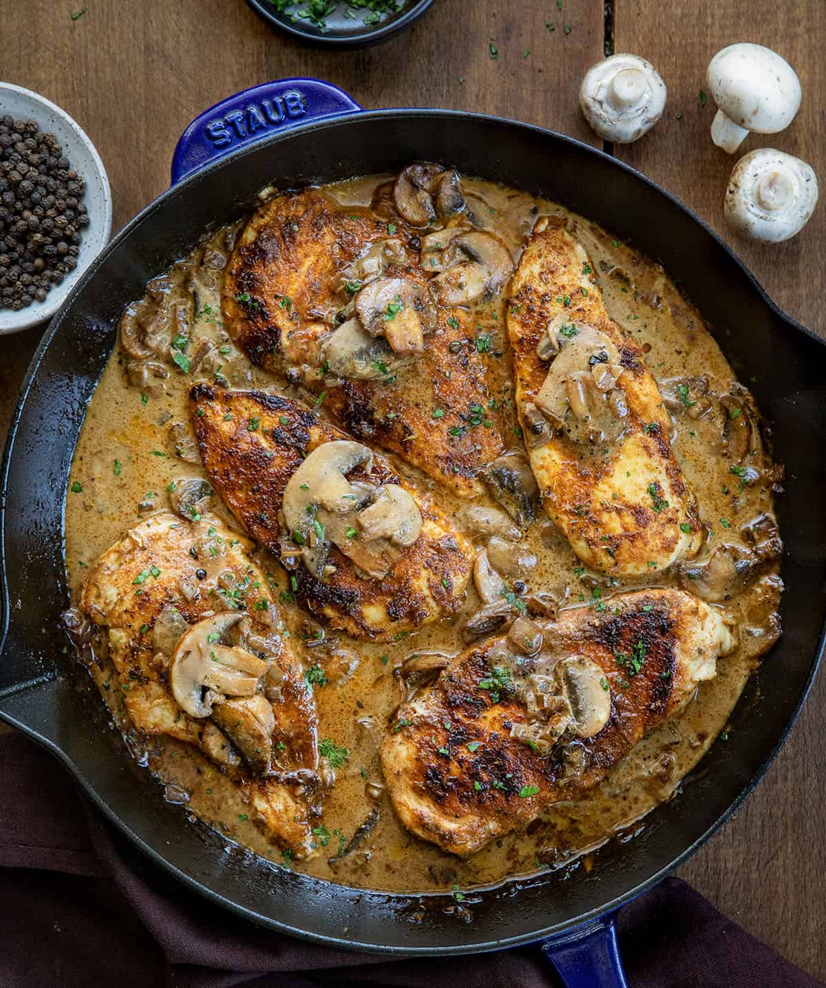 Skillet of Mushroom Peppercorn Chicken on a wooden table with peppercorns and mushrooms Skillet of Mushroom Peppercorn Chicken on a wooden table with peppercorns and mushrooms.