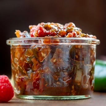 Jar of Raspberry Jalapeño Bacon Jam on a wooden table with fresh raspberry and jalapeno.