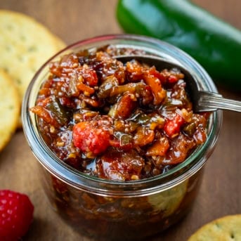 Looking into a jar of Raspberry Jalapeño Bacon Jam with a spoon in it with fresh jalapeno and raspberry.