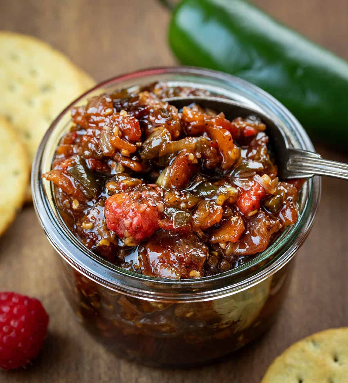 Looking into a jar of Raspberry Jalapeño Bacon Jam with a spoon in it with fresh jalapeno and raspberry Looking into a jar of Raspberry Jalapeño Bacon Jam with a spoon in it with fresh jalapeno and raspberry.