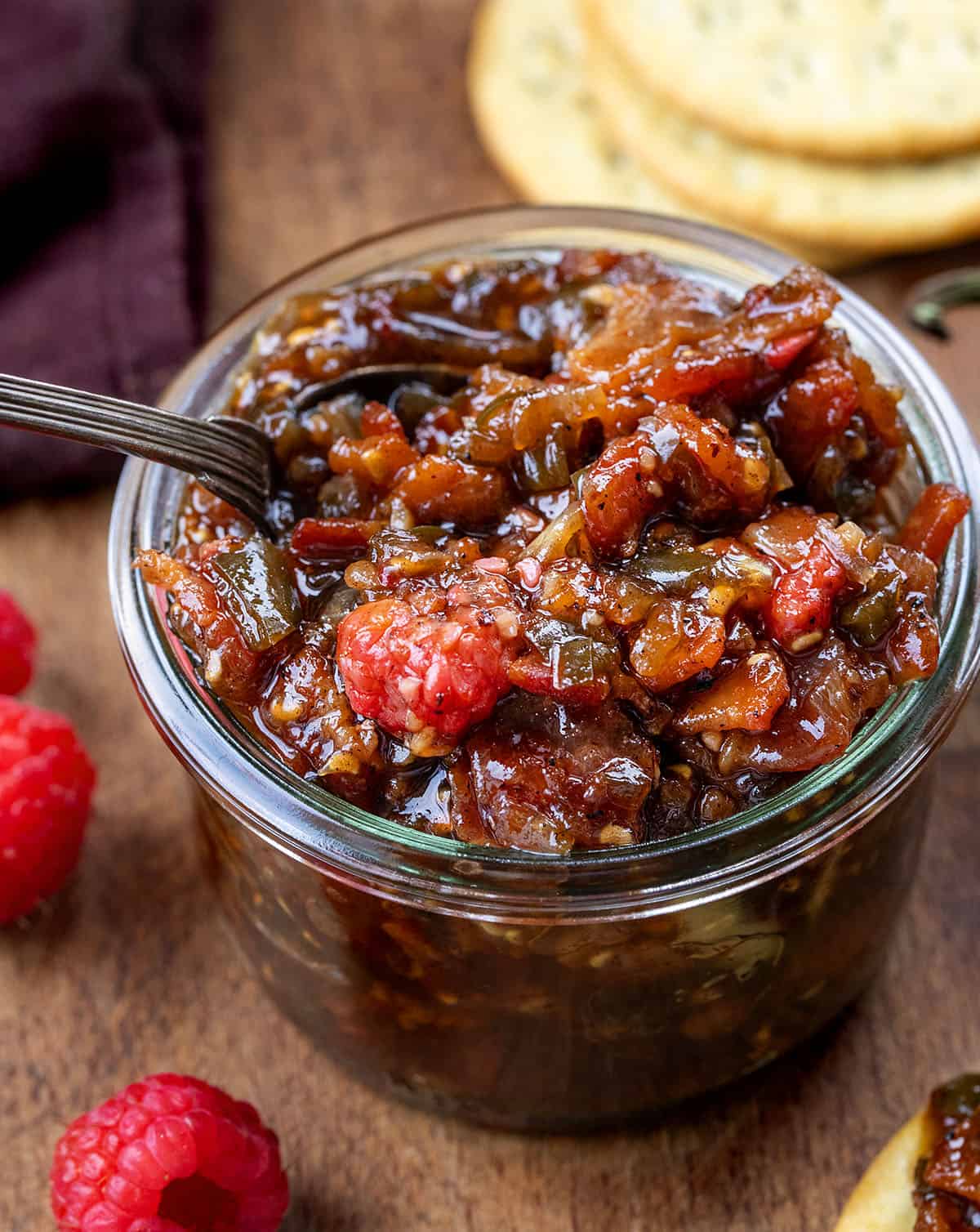 Jar of Raspberry Jalapeño Bacon Jam with a spoon in it on a wooden table with jalapeno and raspberry Jar of Raspberry Jalapeño Bacon Jam with a spoon in it on a wooden table with jalapeno and raspberry.
