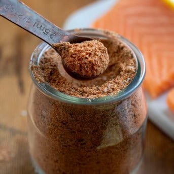 Measuring spoon in a jar of Salmon Seasoning next to some fresh salmon.